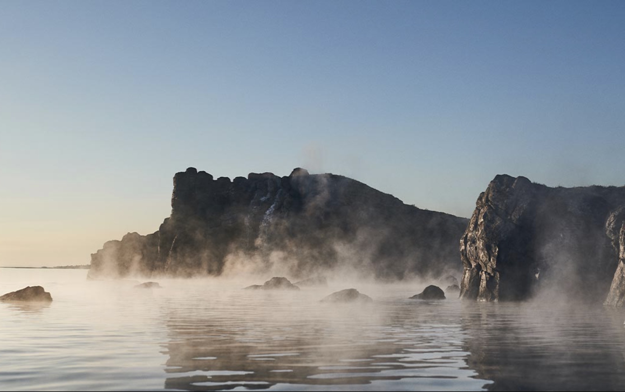 Steaming geothermal waters at Sky Lagoon in Kópavogur with dark rock formations and ocean views.