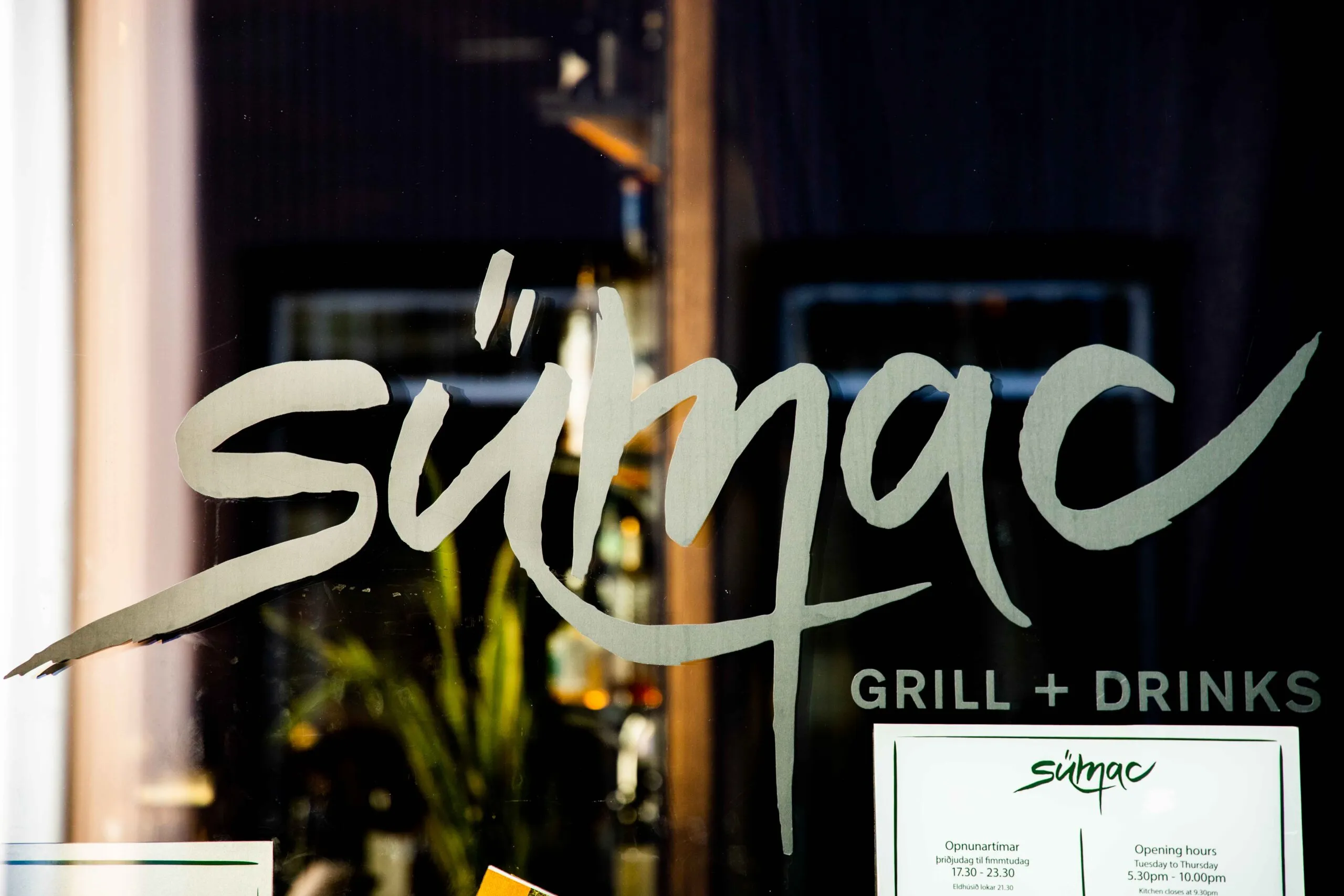 Close-up of Sumac Grill + Drinks logo and opening hours on the window in Reykjavik.