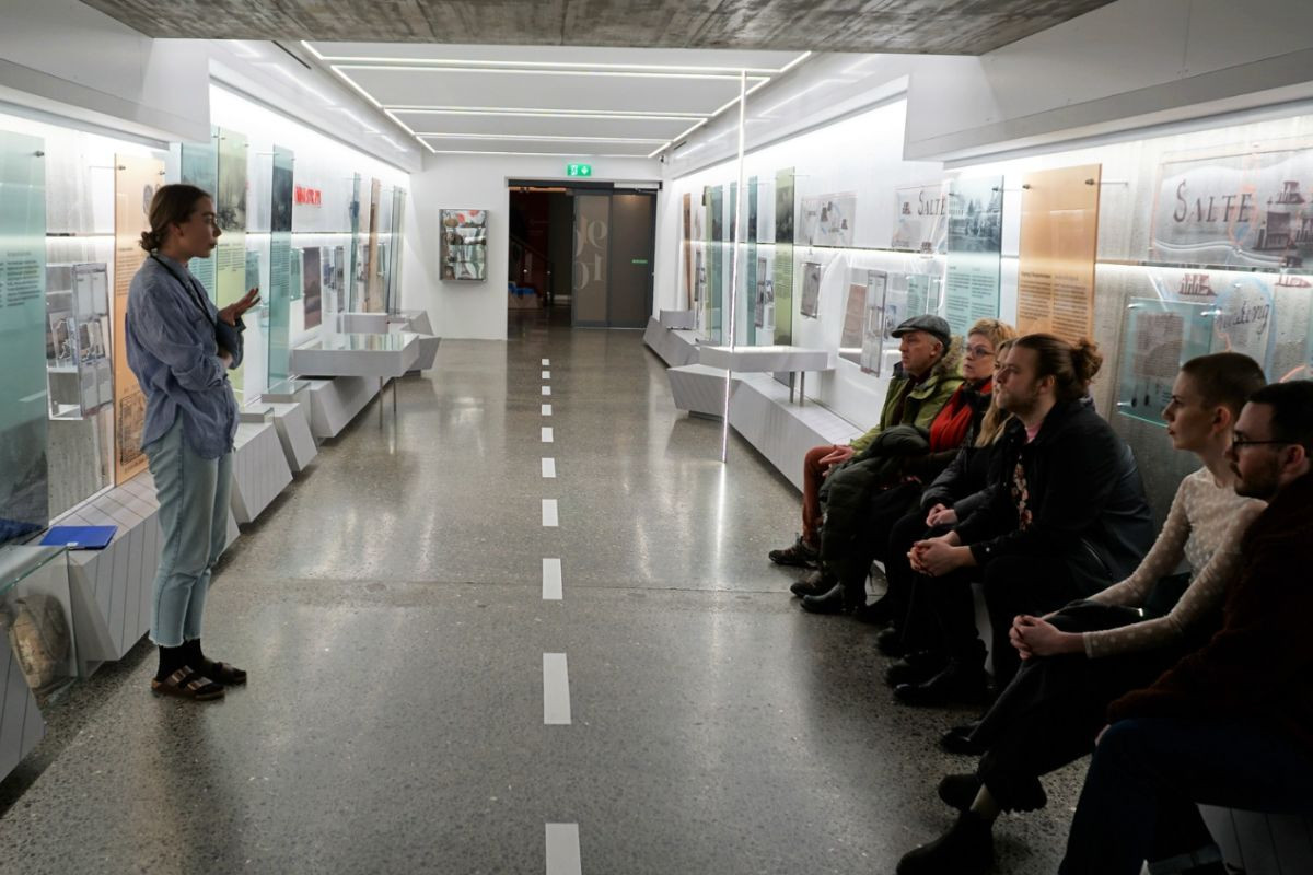 Interior view of The Settlement Exhibition gallery with a tour guide and seated visitors.