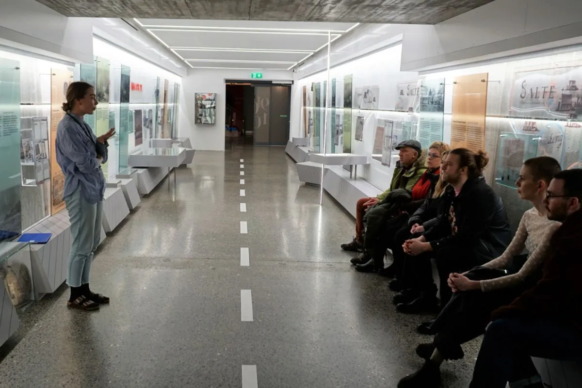 Interior view of The Settlement Exhibition gallery with a tour guide and seated visitors.