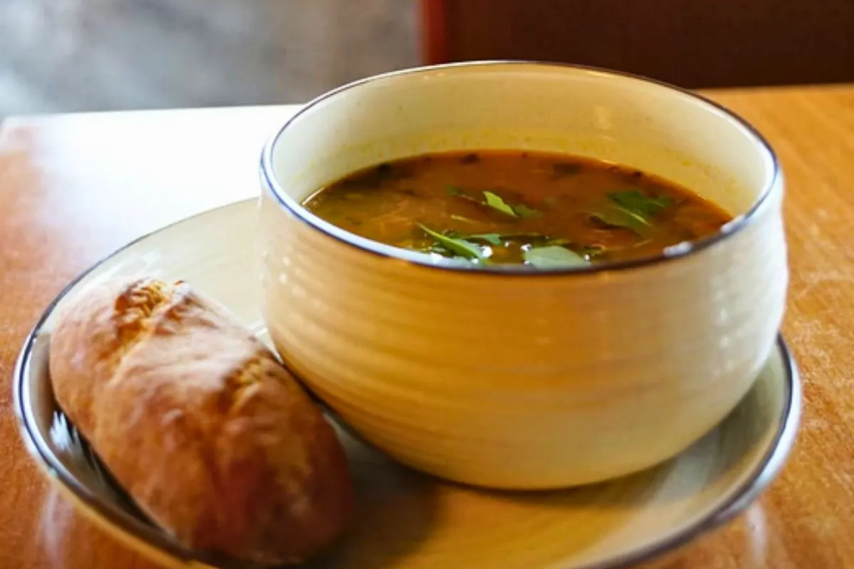A bowl of hot vegetable soup served with a fresh bread roll on a wooden table.