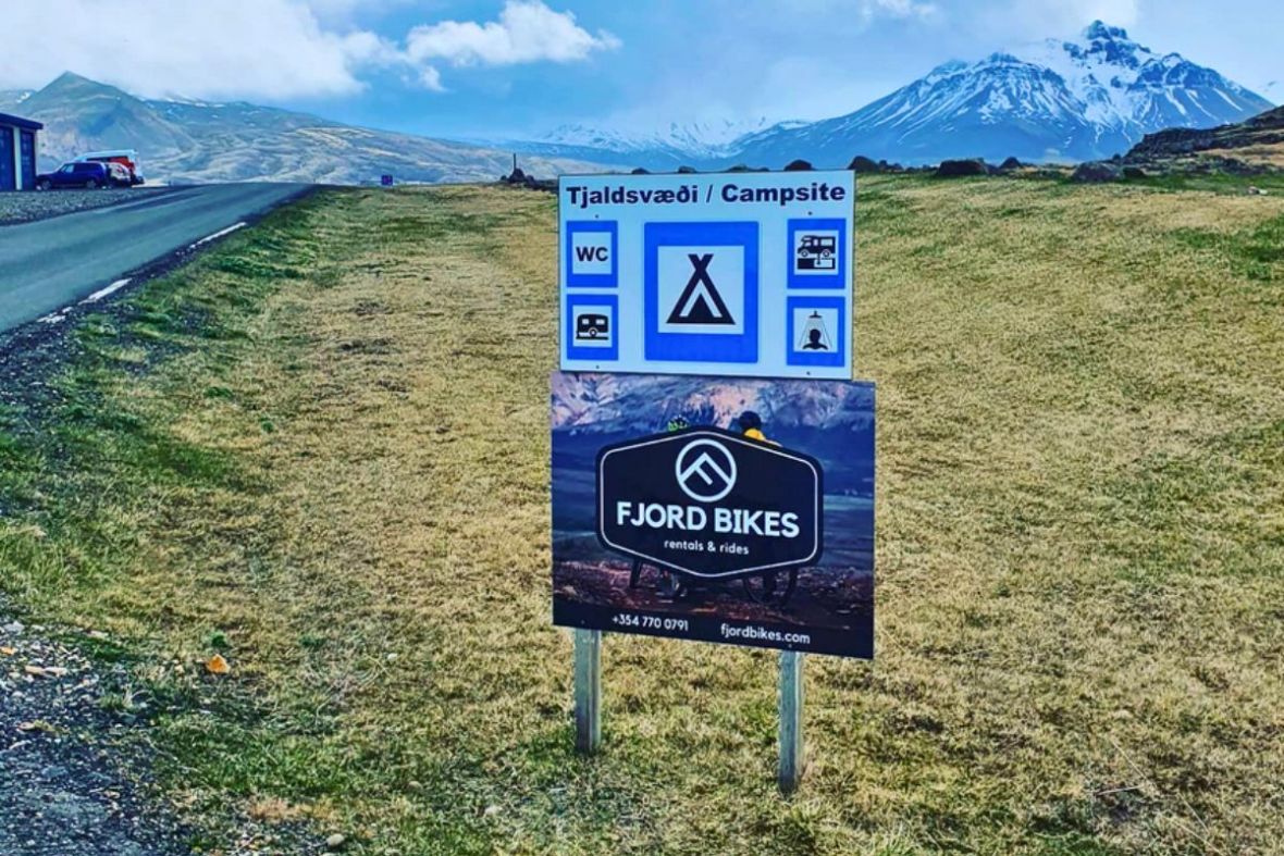 A roadside Fjord Bikes rental sign next to a campsite sign in Borgarfjörður Eystri, East Iceland.