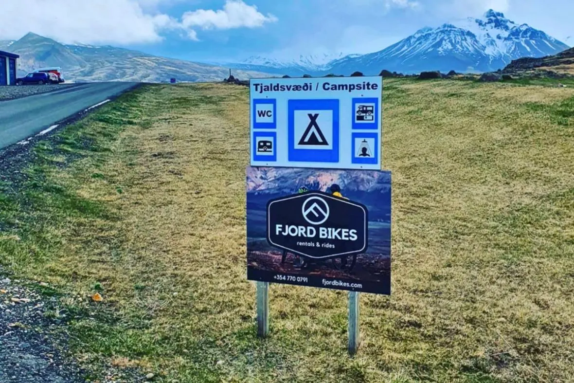 A roadside Fjord Bikes rental sign next to a campsite sign in Borgarfjörður Eystri, East Iceland.