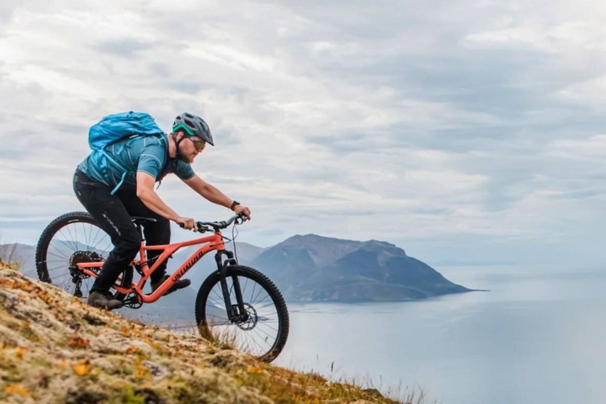 A man riding a Specialized mountain bike down a steep, mossy hill overlooking an Icelandic fjord.