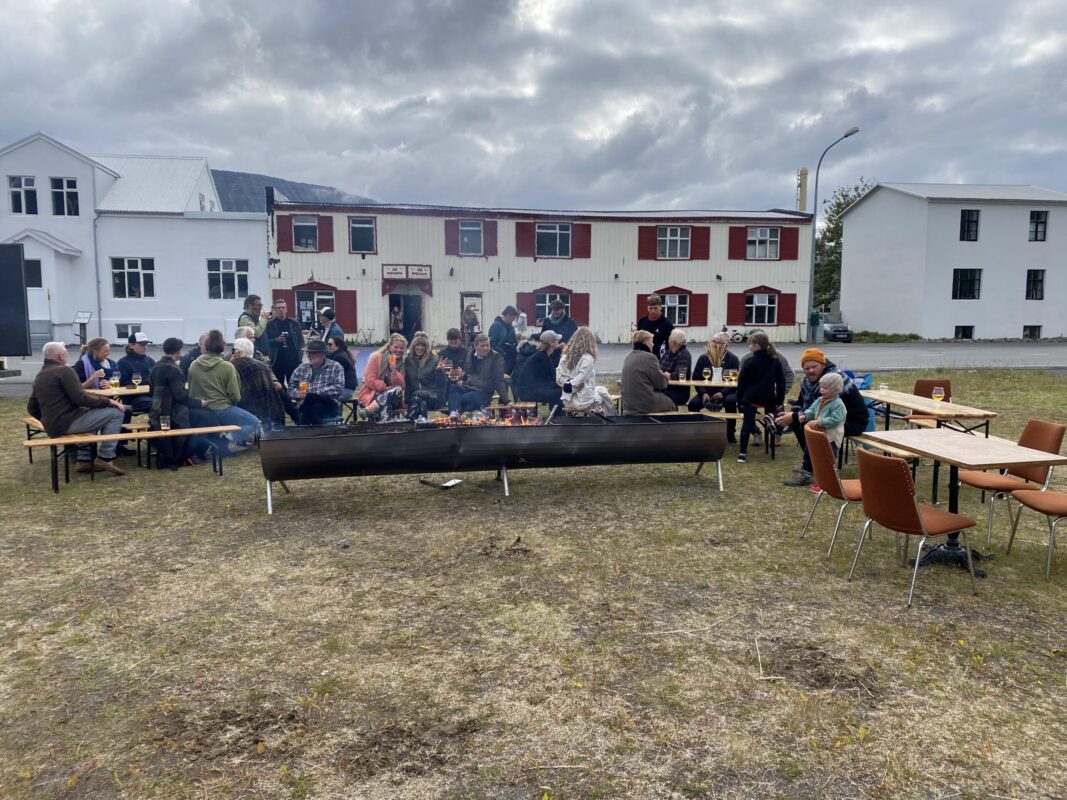 A large group of people sitting at outdoor tables around a long fire pit outside Vagninn in Flateyri.