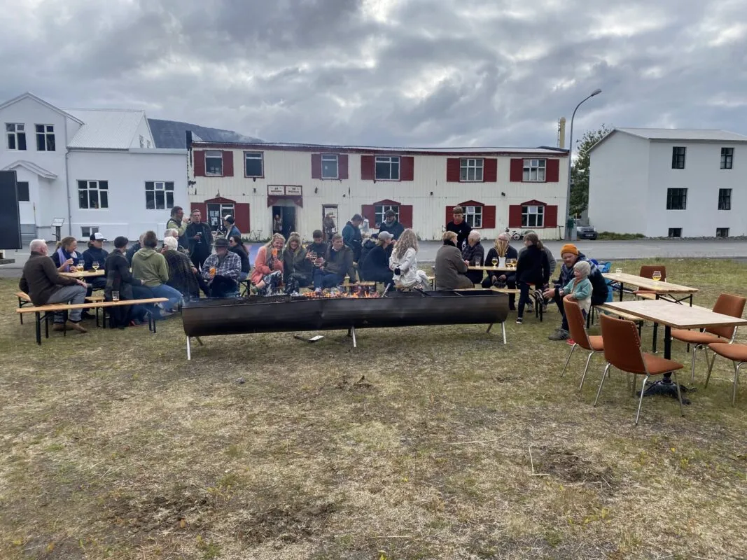 A large group of people sitting at outdoor tables around a long fire pit outside Vagninn in Flateyri.