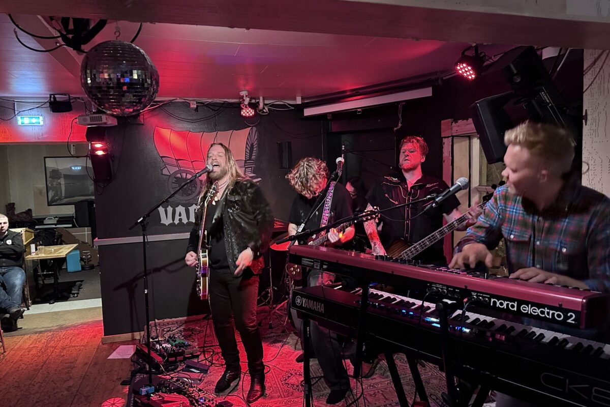 A live rock band with a singer, guitarists, and keyboard player performing on stage under red lights at Vagninn pub.