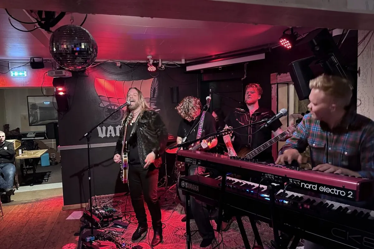 A live rock band with a singer, guitarists, and keyboard player performing on stage under red lights at Vagninn pub.