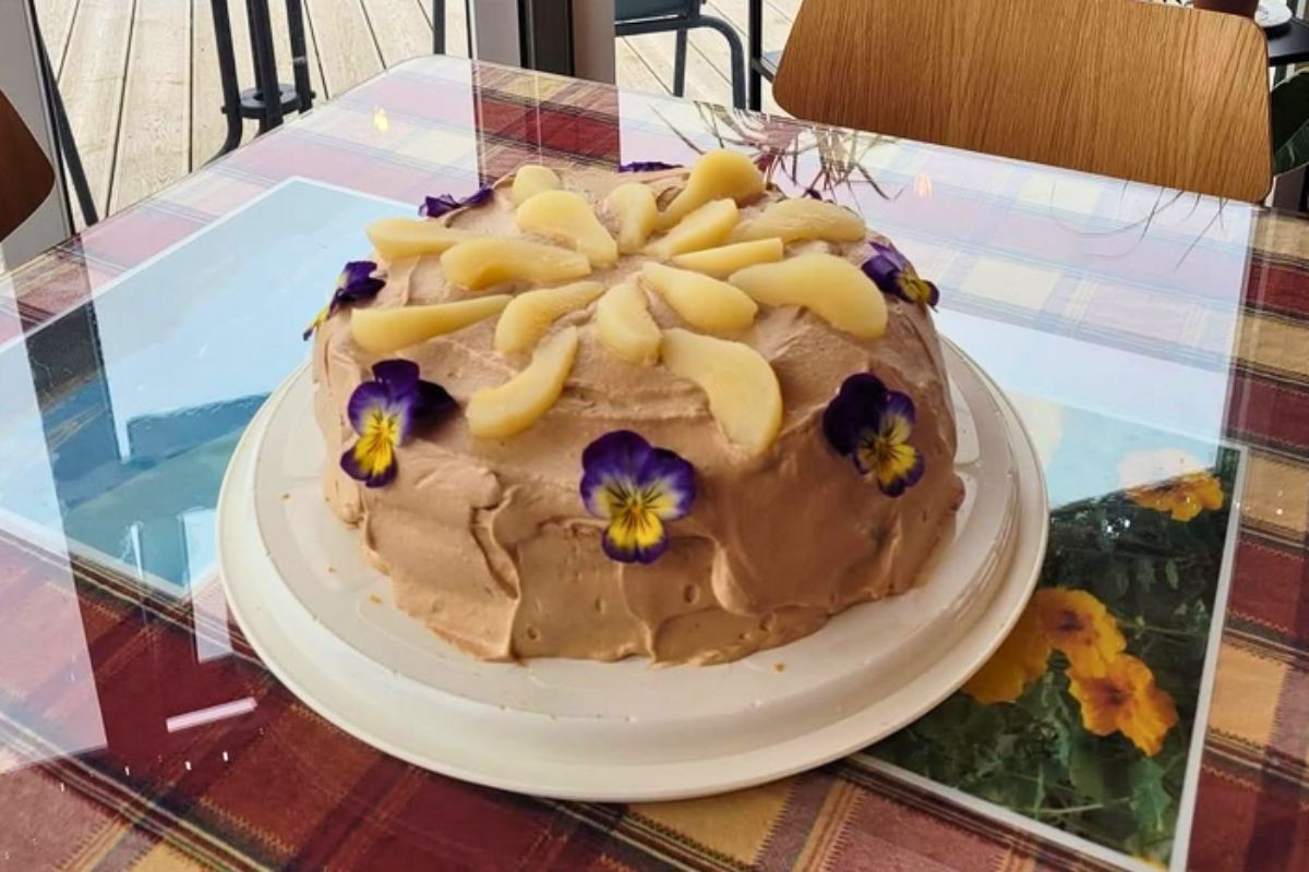 A frosted cake decorated with sliced fruit and purple pansy flowers on a plaid tablecloth.