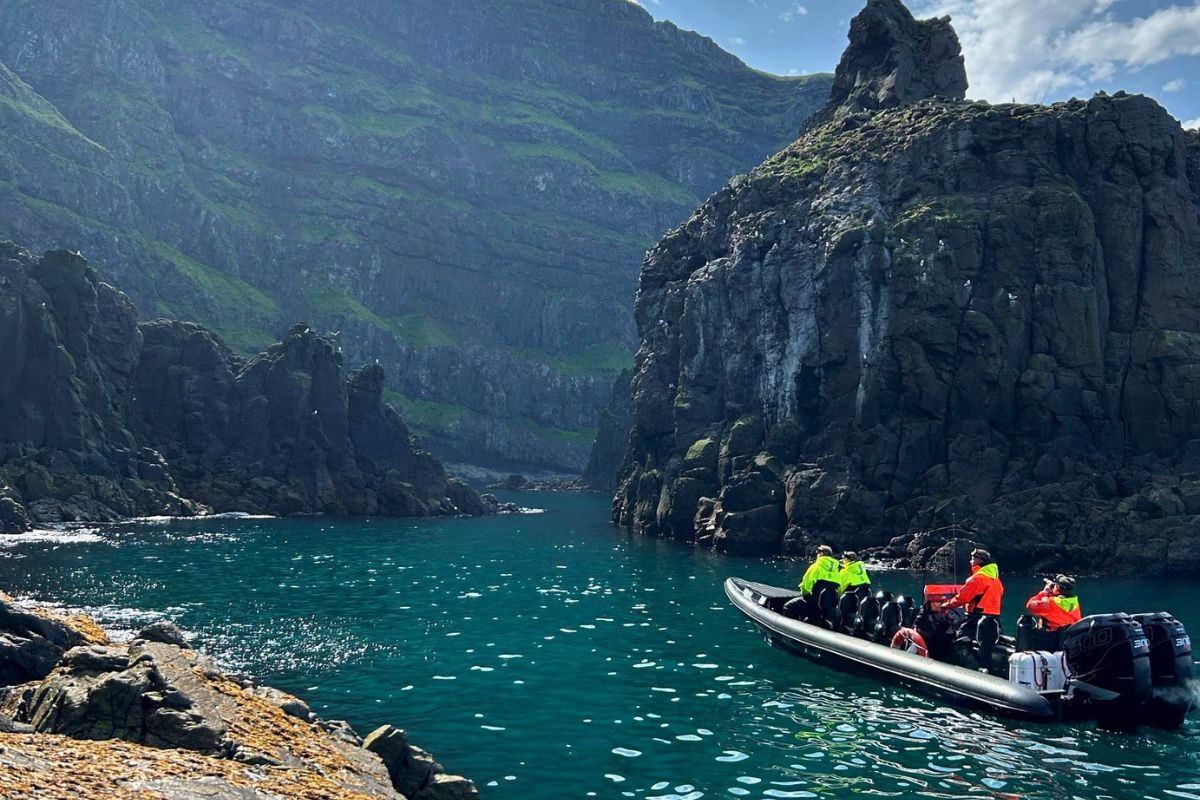 A RIB boat cruising through a narrow turquoise sea channel surrounded by massive towering volcanic cliffs.
