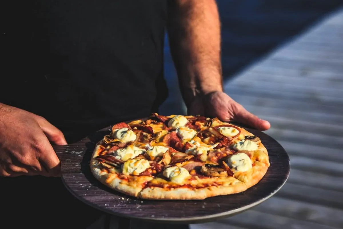 A person holding a freshly baked pizza topped with cream cheese on a wooden board at The Bait Shack (Beituskúrinn) in East Iceland.