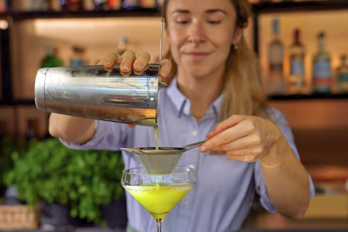 A bartender straining a bright yellow signature cocktail into a coupe glass at Friðheima bar.