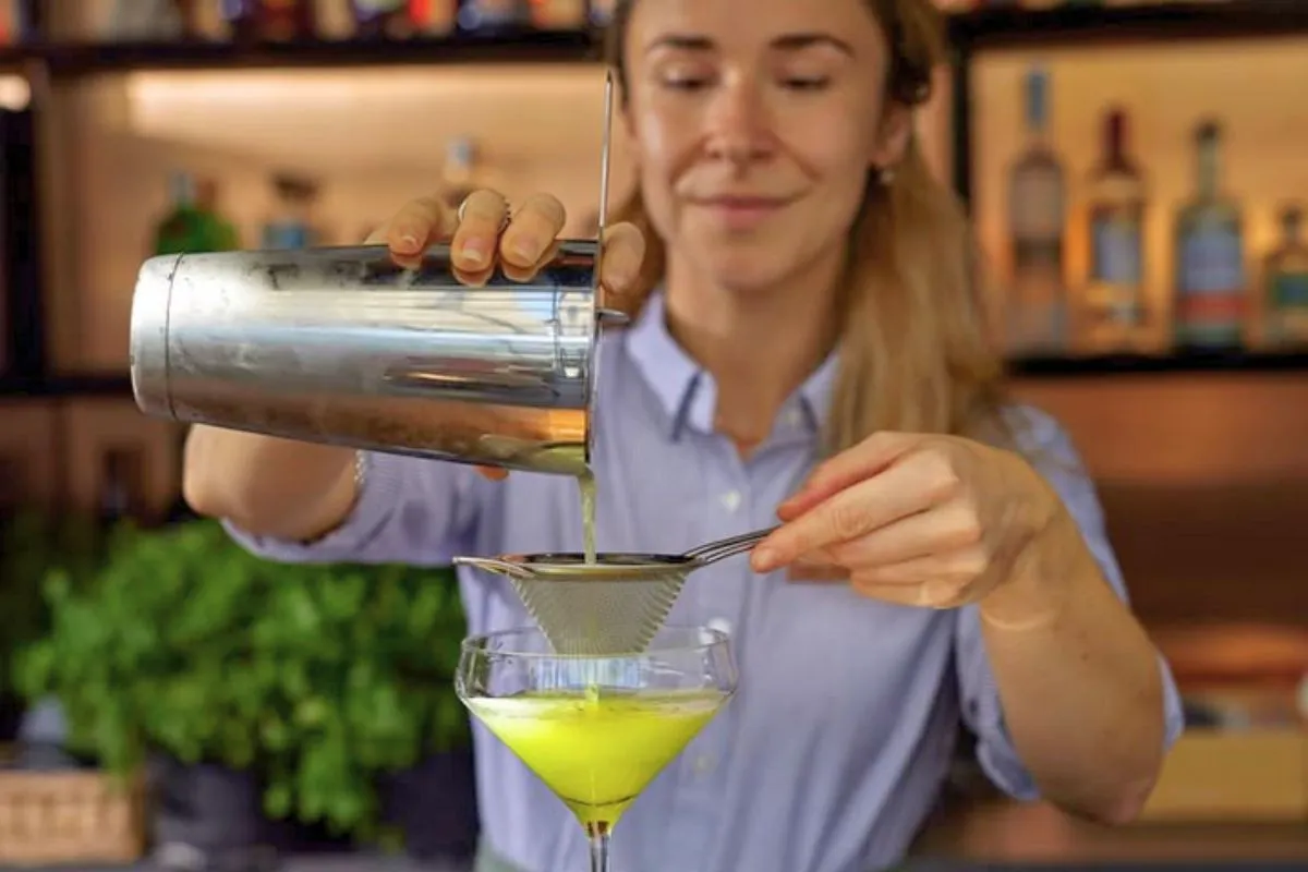 A bartender straining a bright yellow signature cocktail into a coupe glass at Friðheima bar.
